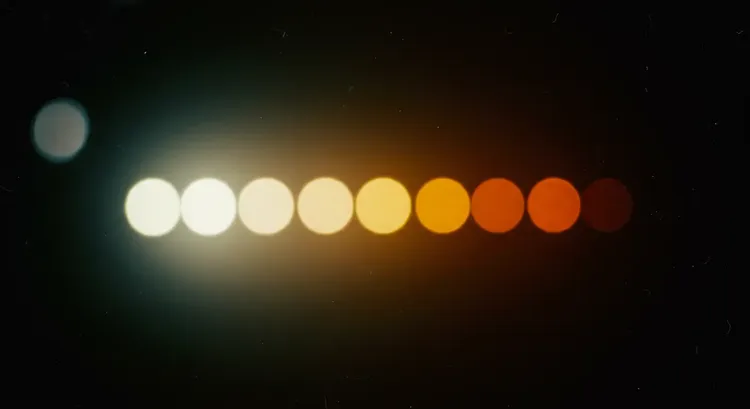 A close-up of an incandescent bulb transitioning from bright white to a warm yellowish glow due to dimming, illustrating the impact of voltage reduction on filament temperature